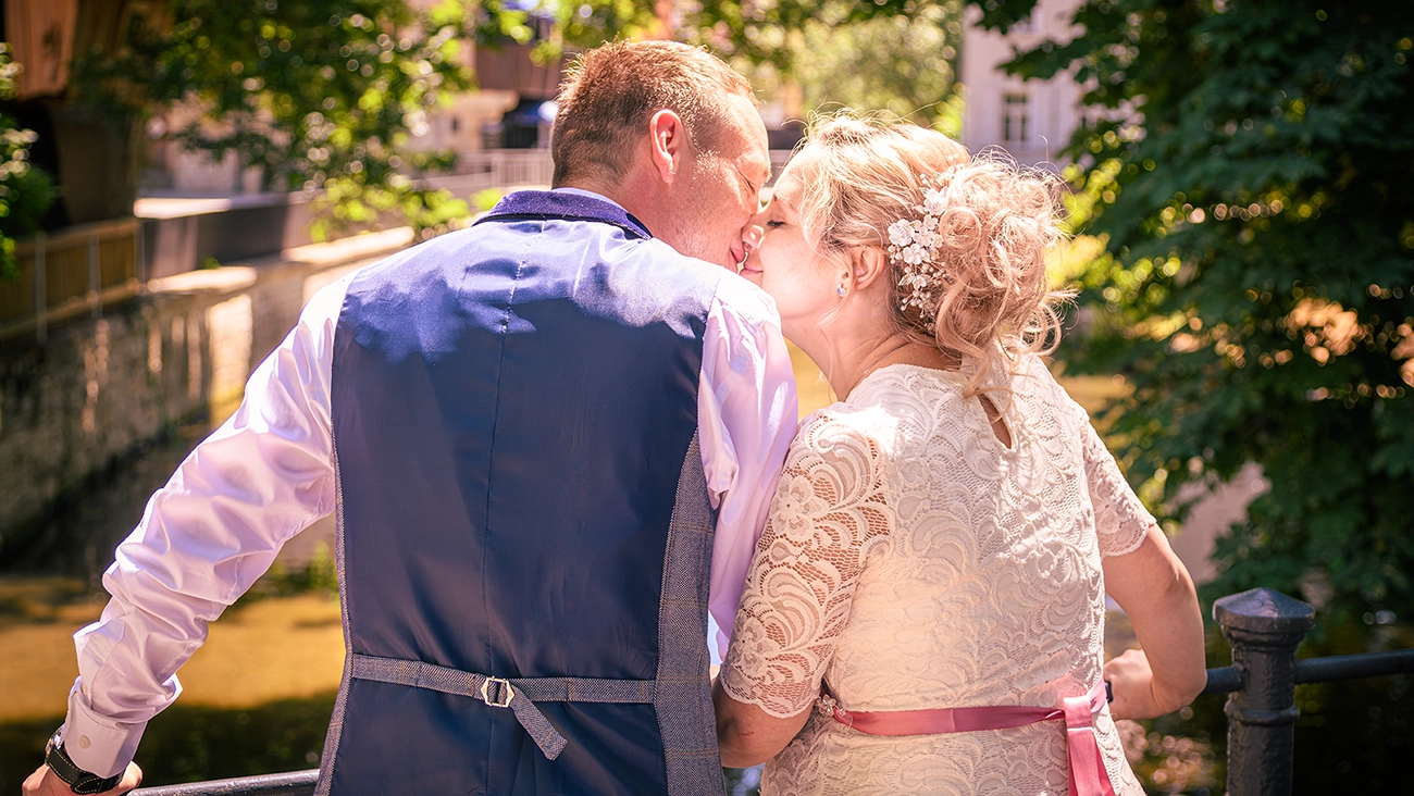 Heiraten in Erfurt: Krämerbrücke als Hochzeitskulisse – M. Schalansky Brautpaar küsst sich innig auf der Erfurter Krämerbrücke, von hinten fotografiert. Die Braut trägt ein Spitzenkleid mit rosa Band, der Bräutigam eine blaue Weste. Im Hintergrund ist die charakteristische Brückenarchitektur zu sehen. Fotografiert von Michael Schalansky.